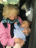 An elderly person is holding a baby in their arms inside a car. Both appear to be engaged in a gentle interaction, with the baby looking up at the elder.