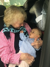 An elderly person is holding a baby in their arms inside a car. Both appear to be engaged in a gentle interaction, with the baby looking up at the elder.