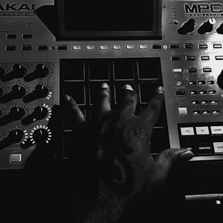 A hand is pressing the pads of a music production controller, surrounded by various knobs and buttons. The dim lighting creates a contrast, highlighting the texture of the hand and the equipment.