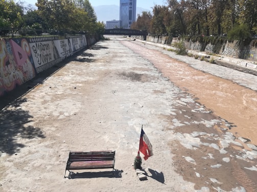 A dried riverbed with graffiti-covered walls on the left side and a small stream of muddy water on the right. A bench and a flag are placed on the dried riverbed under bright sunlight, with overgrown greenery in the background.