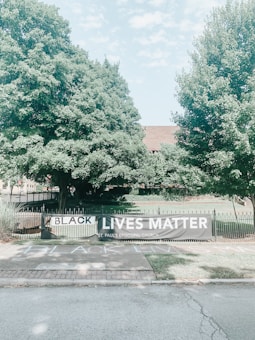 A large banner with the words 'Black Lives Matter' is displayed on a fence in front of a church. The surrounding area features lush green trees and grass, creating a peaceful, natural setting. The sidewalk in front also has a chalk message with the same words.