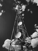 A tall metal communication tower reaching into the sky, equipped with several large satellite dishes. The structure stands against a backdrop of scattered, fluffy clouds in a predominantly dark sky.