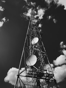 A tall metal communication tower reaching into the sky, equipped with several large satellite dishes. The structure stands against a backdrop of scattered, fluffy clouds in a predominantly dark sky.