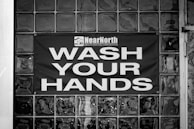 A black and white image displaying a glass block wall with a sign reading 'Wash Your Hands' in large, bold letters. Above the text, there is a logo and the words 'Near North Health Service Corporation.' The sign is centered on the wall, emphasizing cleanliness and hygiene.
