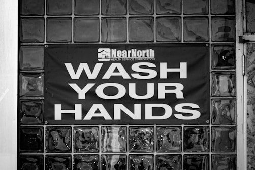 A black and white image displaying a glass block wall with a sign reading 'Wash Your Hands' in large, bold letters. Above the text, there is a logo and the words 'Near North Health Service Corporation.' The sign is centered on the wall, emphasizing cleanliness and hygiene.