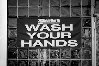 A black and white image displaying a glass block wall with a sign reading 'Wash Your Hands' in large, bold letters. Above the text, there is a logo and the words 'Near North Health Service Corporation.' The sign is centered on the wall, emphasizing cleanliness and hygiene.