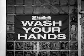 A black and white image displaying a glass block wall with a sign reading 'Wash Your Hands' in large, bold letters. Above the text, there is a logo and the words 'Near North Health Service Corporation.' The sign is centered on the wall, emphasizing cleanliness and hygiene.