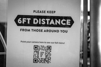 A black and white sign instructs people to maintain a distance of 6 feet from those around them. There is a QR code at the bottom for accessing a full menu with a directive to point a camera at it.