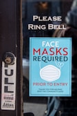 A sign on a glass door with instructions to ring the bell and a notice stating that face masks are required prior to entry. The sign is blue with white text and includes an image of a face mask. There is also a smaller sticker for a door repair service on the door.