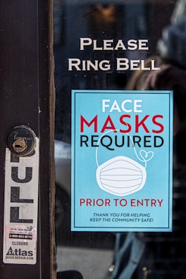 A sign on a glass door with instructions to ring the bell and a notice stating that face masks are required prior to entry. The sign is blue with white text and includes an image of a face mask. There is also a smaller sticker for a door repair service on the door.