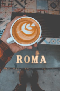A person is holding a cup of coffee with latte art on the surface. The background includes colorful patterned tiles and the word 'ROMA' on the ground near the person's feet.