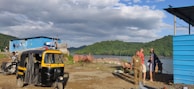 A scenic lakeside area with lush green hills in the background. In the foreground, there is a yellow and black auto rickshaw parked on a dirt path. Several people are gathered near a group of blue corrugated metal shacks beside a large body of water, with some clothing hanging to dry. The sky is partly cloudy, creating a peaceful and relaxed atmosphere.