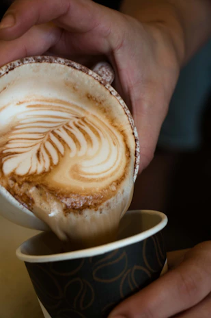 Close-up of hands holding a coffee cup with latte art, surrounded by coffee beans and a notebook.