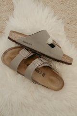 Two beige Birkenstock sandals are placed casually on a fluffy white rug. The sandals have a simple, comfortable design with leather straps and metallic buckles.