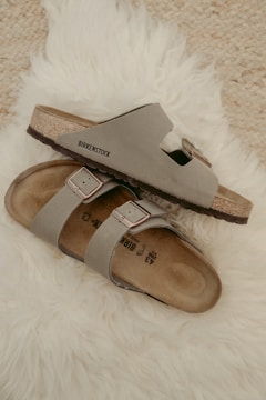 Two beige Birkenstock sandals are placed casually on a fluffy white rug. The sandals have a simple, comfortable design with leather straps and metallic buckles.