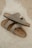 Two beige Birkenstock sandals are placed casually on a fluffy white rug. The sandals have a simple, comfortable design with leather straps and metallic buckles.