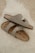 Two beige Birkenstock sandals are placed casually on a fluffy white rug. The sandals have a simple, comfortable design with leather straps and metallic buckles.