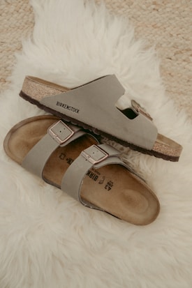 Two beige Birkenstock sandals are placed casually on a fluffy white rug. The sandals have a simple, comfortable design with leather straps and metallic buckles.