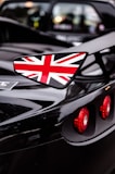 A close-up shot of a sports car highlighting its glossy black finish. The vehicle prominently features a side wing mirror adorned with the Union Jack, and the red circular taillights stand out against the sleek surface.
