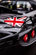 A close-up shot of a sports car highlighting its glossy black finish. The vehicle prominently features a side wing mirror adorned with the Union Jack, and the red circular taillights stand out against the sleek surface.