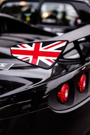 A close-up shot of a sports car highlighting its glossy black finish. The vehicle prominently features a side wing mirror adorned with the Union Jack, and the red circular taillights stand out against the sleek surface.