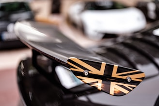A close-up of a car wing with a Union Jack design in black and gold. The background is blurred, showing other cars in a parking lot.