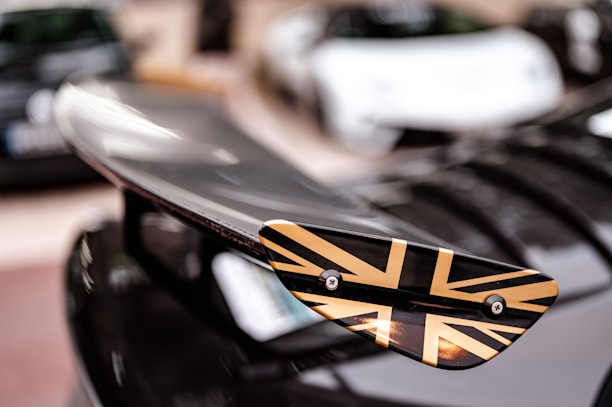 A close-up of a car wing with a Union Jack design in black and gold. The background is blurred, showing other cars in a parking lot.