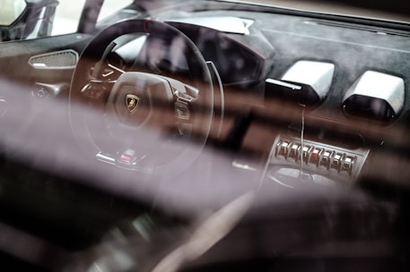 A luxury car interior showcases a sleek steering wheel with a prominent logo, surrounded by high-end materials and advanced controls. The dashboard features modern design elements and metallic accents, emphasizing a sense of sophistication and speed.