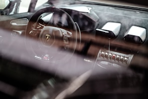 A luxury car interior showcases a sleek steering wheel with a prominent logo, surrounded by high-end materials and advanced controls. The dashboard features modern design elements and metallic accents, emphasizing a sense of sophistication and speed.