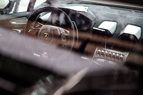 A luxury car interior showcases a sleek steering wheel with a prominent logo, surrounded by high-end materials and advanced controls. The dashboard features modern design elements and metallic accents, emphasizing a sense of sophistication and speed.