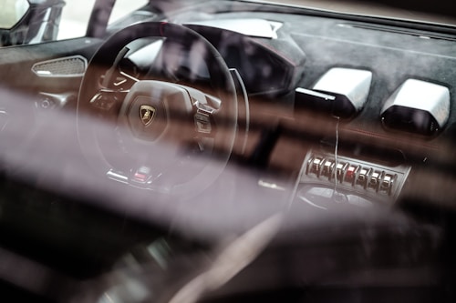 A luxury car interior showcases a sleek steering wheel with a prominent logo, surrounded by high-end materials and advanced controls. The dashboard features modern design elements and metallic accents, emphasizing a sense of sophistication and speed.