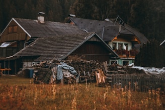 A rustic house surrounded by a wooded area, featuring a dark brown wooden exterior and a steeply pitched roof. In front of the house, a large pile of chopped firewood is neatly stacked, covered partially by tarps. The chimney is visible on the roof, and the balcony of the house is adorned with potted flowers. The overall scene is peaceful, set in a rural environment.