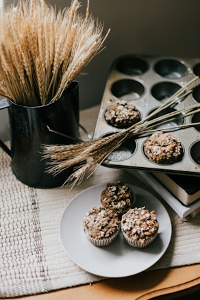 Muffins with a crumbly topping are placed on a white plate, with additional muffins still in a baking tin. A black jug containing wheat stalks is positioned nearby, creating a rustic and cozy atmosphere.