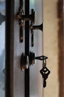 A close-up view of a door with two metallic locks. One of the locks contains a key with a simple design, slightly bent at an angle. The door appears to be made of dark wood, and there is a muted, blurry background visible through a gap in the door.