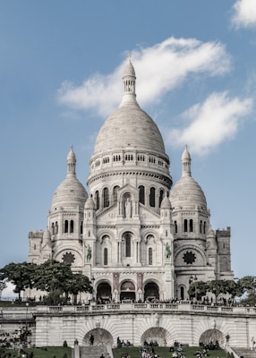 A large, ornate basilica featuring a central dome and two smaller domes flanking it. The structure is built in a Romanesque-Byzantine style and is made of white stone. There are green statues of knights on horseback on either side of the central entrance. The building is set against a blue sky with a few fluffy clouds. In front of the basilica, a green lawn with people walking and sitting creates a lively atmosphere.