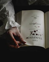 An open book with the name 'Anne Rice' and the title 'The Vampire Lestat' is displayed. A hand with visible blood stains is turning the page. The book and hand are against a dark background, enhancing the mysterious and eerie atmosphere.