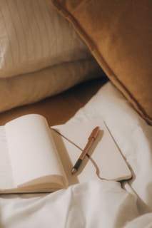 A soft, cozy setting featuring a notebook and pen resting on a bed with white and brown cushions. The scene suggests a personal, intimate moment of writing or reflection.