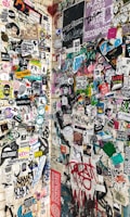 A wall covered with a diverse array of stickers, posters, and graffiti, featuring various colorful illustrations, logos, and texts. The designs range from cartoon characters and artistic expressions to brand and event advertisements. The surface appears to be an urban setting, capturing an eclectic mix of street art and underground culture.