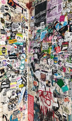 A wall covered with a diverse array of stickers, posters, and graffiti, featuring various colorful illustrations, logos, and texts. The designs range from cartoon characters and artistic expressions to brand and event advertisements. The surface appears to be an urban setting, capturing an eclectic mix of street art and underground culture.