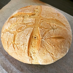 A round, rustic loaf of bread with a crusty exterior and intricate scoring patterns on the top. The bread has a golden-brown color and sits on a piece of parchment paper.