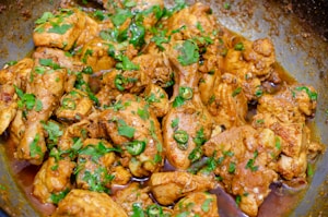 A close-up of cooked chicken pieces garnished with fresh green herbs and sliced green chilies, served in a pan. The chicken appears to be seasoned with spices, giving it a rich, savory look.
