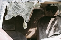 A damaged car interior with a shattered windshield and dusty seats. The broken glass is jagged and partially hanging down, casting shadows inside the vehicle.