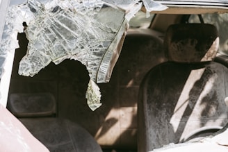 A damaged car interior with a shattered windshield and dusty seats. The broken glass is jagged and partially hanging down, casting shadows inside the vehicle.