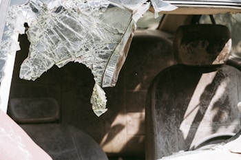 A damaged car interior with a shattered windshield and dusty seats. The broken glass is jagged and partially hanging down, casting shadows inside the vehicle.