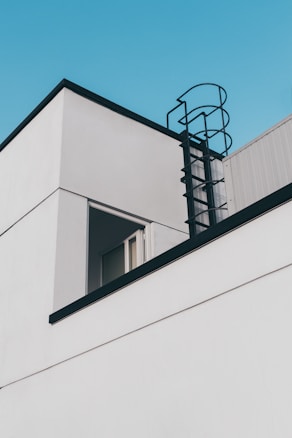 A minimalistic modern building with clean, white walls and a black metal exterior ladder leading to the rooftop is set against a clear blue sky. The corner of the building features an open window, adding to the simplistic architectural design.