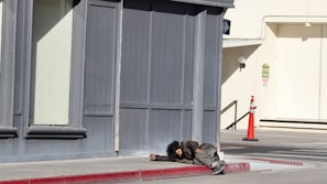 A person is lying on the sidewalk near a corner of a building. The clothing appears worn and disheveled, suggesting a state of homelessness or distress. The surrounding area includes a sidewalk, a red-painted curb, and a distant traffic cone near a building with gray and beige tones.