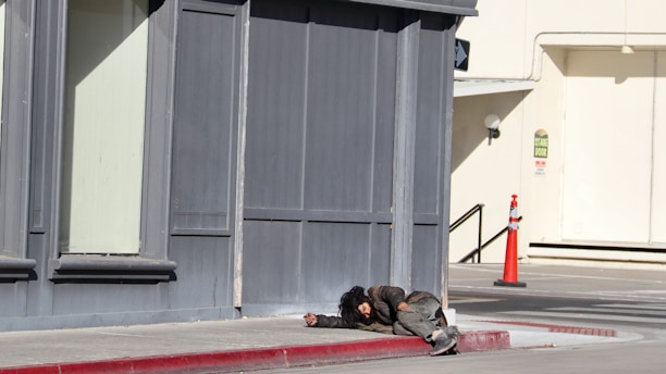 A person is lying on the sidewalk near a corner of a building. The clothing appears worn and disheveled, suggesting a state of homelessness or distress. The surrounding area includes a sidewalk, a red-painted curb, and a distant traffic cone near a building with gray and beige tones.