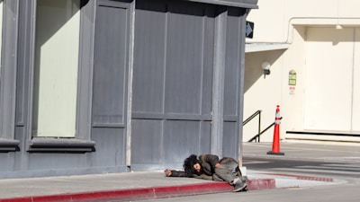 A person is lying on the sidewalk near a corner of a building. The clothing appears worn and disheveled, suggesting a state of homelessness or distress. The surrounding area includes a sidewalk, a red-painted curb, and a distant traffic cone near a building with gray and beige tones.
