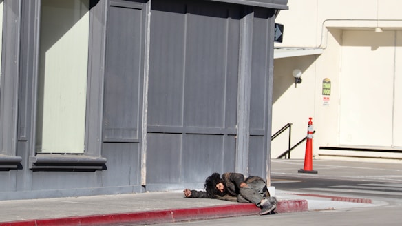 A person is lying on the sidewalk near a corner of a building. The clothing appears worn and disheveled, suggesting a state of homelessness or distress. The surrounding area includes a sidewalk, a red-painted curb, and a distant traffic cone near a building with gray and beige tones.