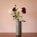 A simple, elegant arrangement of burgundy and white flowers displayed in a tall, cylindrical ceramic vase. The background is a muted, warm beige color, enhancing the natural tones of the flowers and vase. The flowers are fresh, with lush green leaves and long stems.